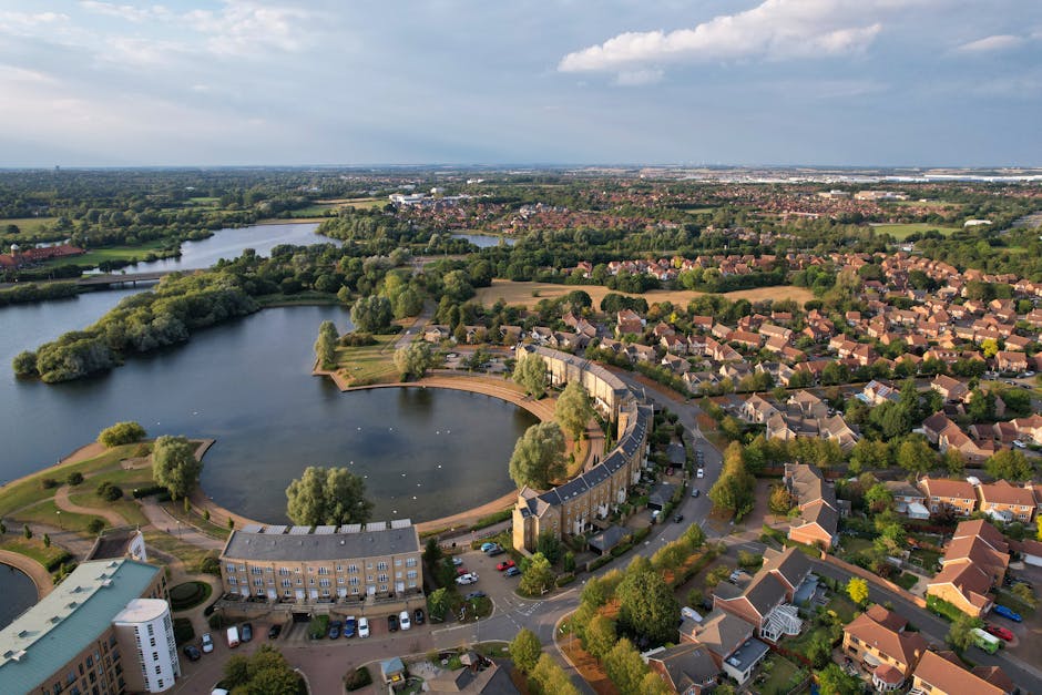 An aerial view of Barnes Common and Pond, showcasing a large, circular pond at the centre surrounded by well-maintained residential houses with terracotta and grey tiled roofs. The pond features a cur