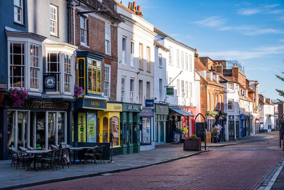 A row of colorful shops and cafes on Barnes High Street, featuring traditional brick and plaster facades with large windows, some decorated with flower baskets and signage. The pavement in front is cl